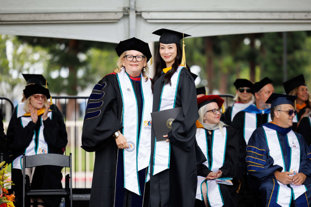 a graduate stands on a stage with a professor, both wearing graduation robes, caps and sashes, smiling and posing together
