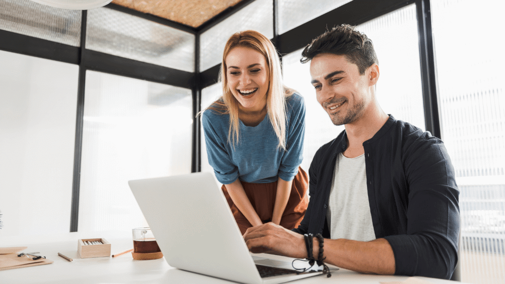 a man sits at a desk in an office setting with an open laptop in front of him, a female looks over his shoulder at the open laptop