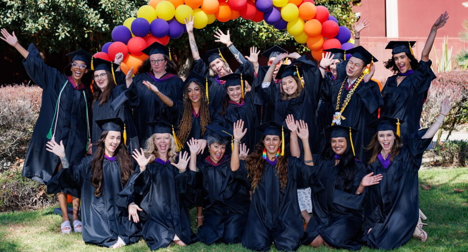 Group of graduates in black robes and caps celebrating under a colorful balloon arch.