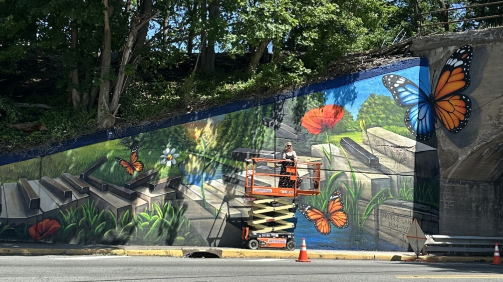 A mural artist on a lift paints a large-scale butterfly and flower garden scene on a concrete wall.