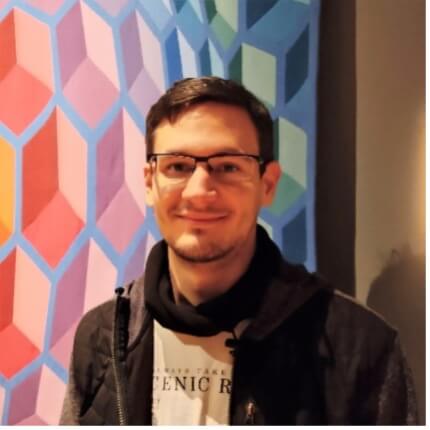 A person wearing glasses and a black hoodie smiles at the camera in front of a colorful geometric.