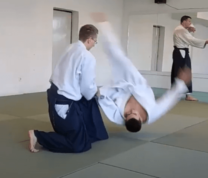 Two people practicing aikido on a mat, with one person being thrown over the other's shoulder.